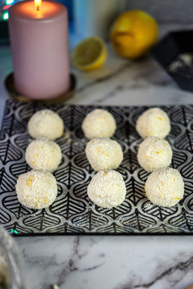 Nine high protein cottage cheese lemon balls on a decorative patterned tray with a lemon and candle in the background.