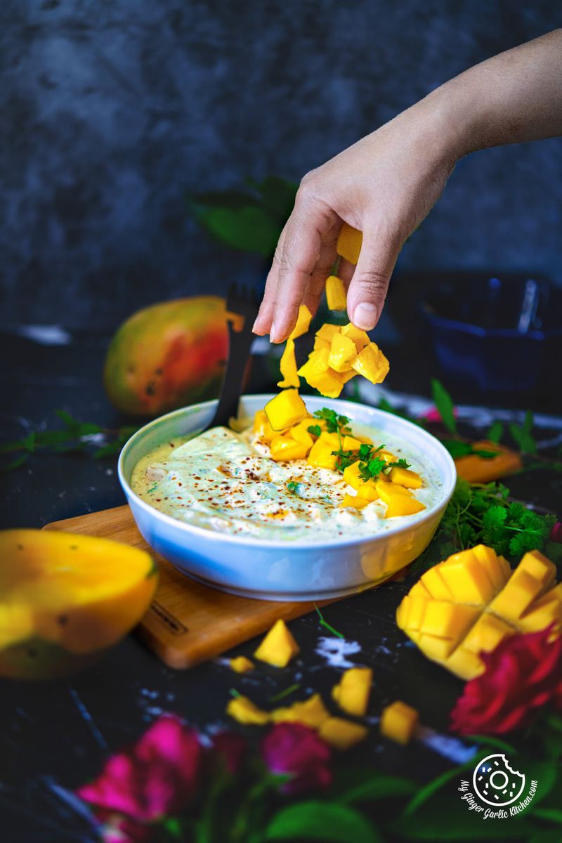 a hand sprinkling mango chunk over a mango raita bowl